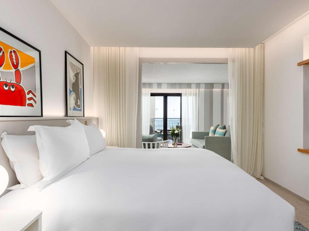 Mūsdienīga viesnīcas istaba ar gultu un mākslas darbiem uz sienām, kas sniedz skatu uz jūru. Interjerā redzamas gaišas krāsas un stilīgas mēbeles.