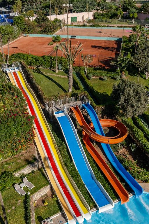 Krāsaini ūdens slidkalniņi, kas atrodas pie baseina, ar blakus esošu tenisa kortu un zaļo ainavu. Ideāla vieta izklaidei un atpūtai.