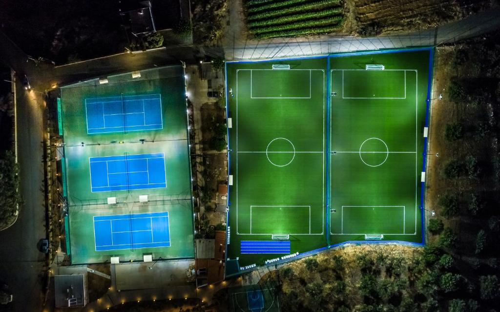 Apskatāma nakts ainava ar vairākiem sporta laukumiem, tostarp tenisa kortiem un futbola laukumiem, apgaismotiem zaļajā krāsā.
