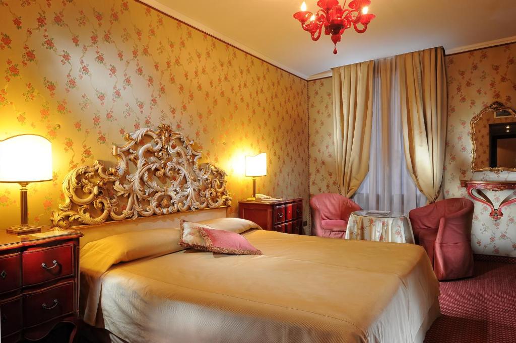 Elegants guļamistabas interjers ar ziedu rakstu tapetēm, greznu gultas galvgalu un komfortablām mēbelēm. Siltas krāsas un mīkstais apgaismojums rada mājīgu atmosfēru.