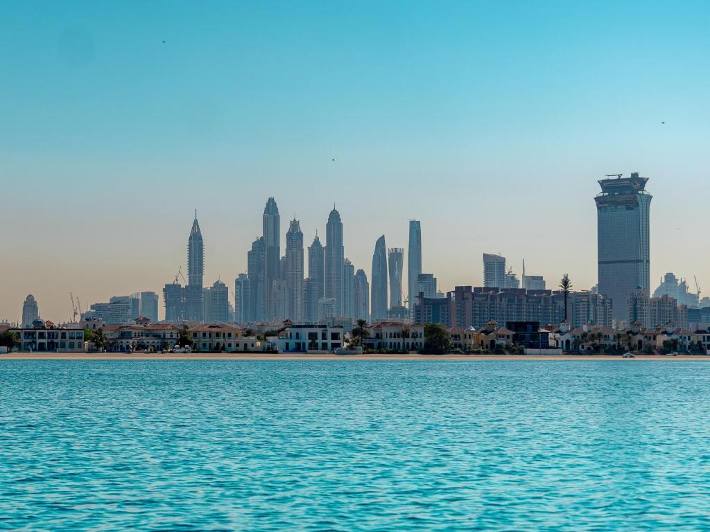 Pilsētas ainava ar moderniem debesskrāpjiem un skaistu ūdens virsmu priekšplānā, radot dinamisku un mūsdienīgu atmosfēru.