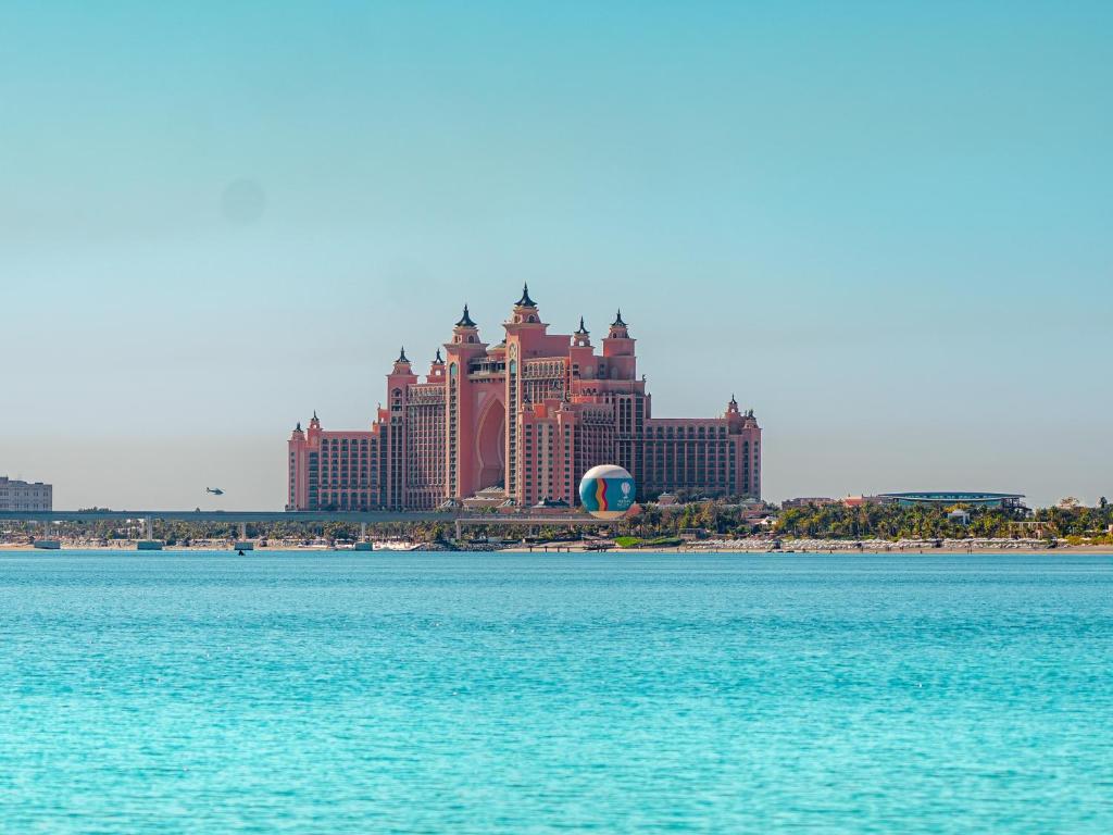 Lielais rozā viesnīcas komplekss pie ūdens, ar skaidru zilu debesi fonā. Ideāla vieta atpūtai un izklaidei.