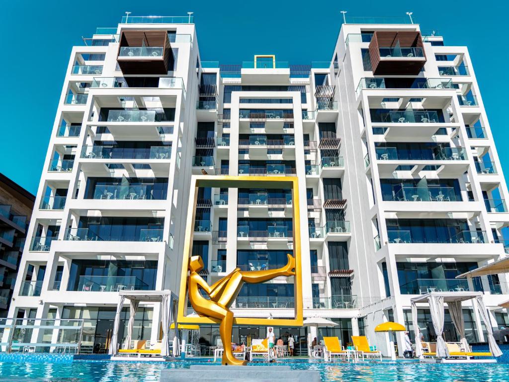 Moderns viesnīcas komplekss ar stikla balkoniem un peldbaseinu, centrā redzama zelta skulptūra, kas simbolizē atpūtu. Klaras debesis papildina mūsdienu arhitektūru.