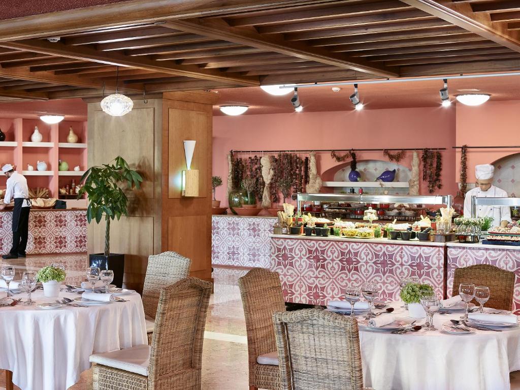 Elegants restorāns ar bagātīgu bufeti, kurā redzami šefpavāri un rūpīgi sakārtoti galdi. Siltas krāsas un dabīgi materiāli rada patīkamu atmosfēru.
