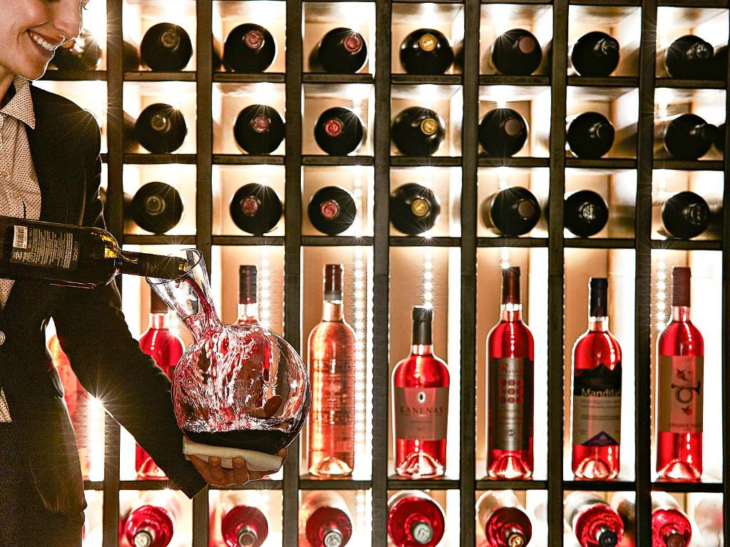 Sommelier ielej sarkanvīnu dekantērā, kam aizmugurē redzams elegants vīna plaukts ar dažādām pudelēm.