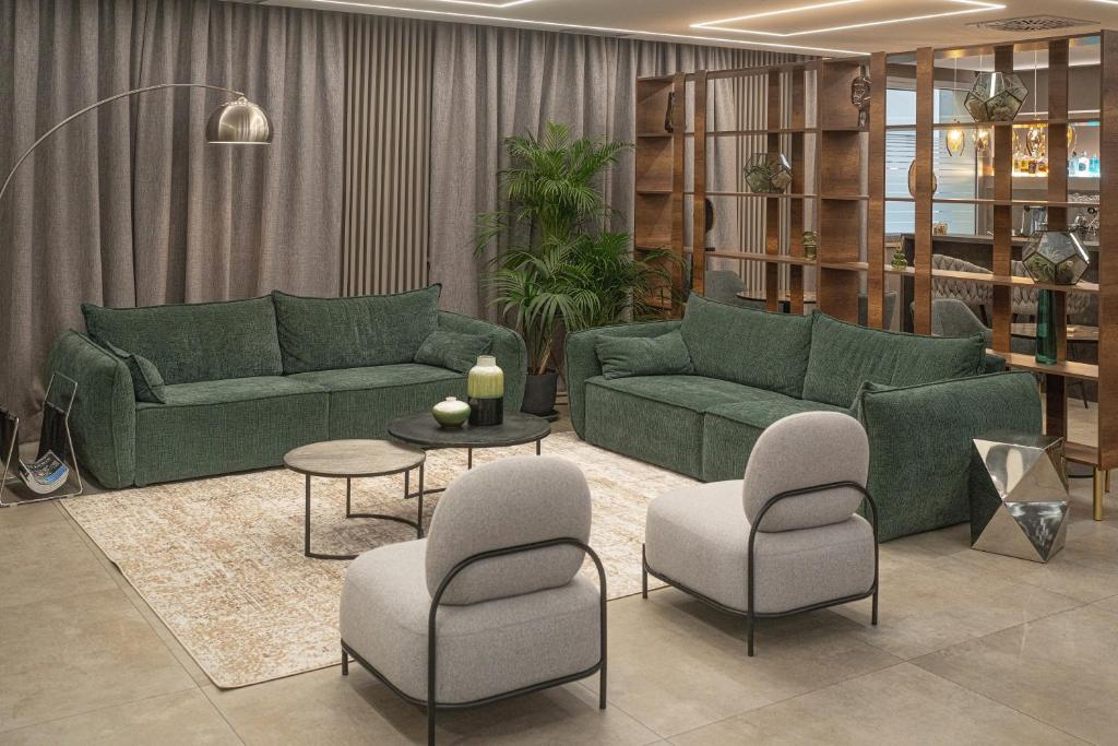 Mūsdienīgs viesistabas interjers ar zaļām dīvāniem, pelēkām krēslām un elegantu kafijas galdiņu. Telpā ir arī dekoratīvi elementi un apgaismojums, kas rada mājīgu atmosfēru.