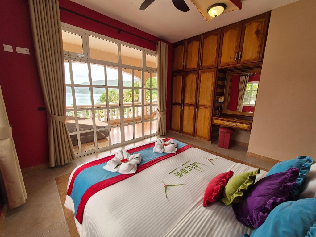 Mūsdienīga guļamistaba ar plašiem logiem, kas paver skatu uz dabu. Gultā ir krāsainas spilvenu dekorācijas un izkārtoti dvieļi, radot siltu un viesmīlīgu atmosfēru.