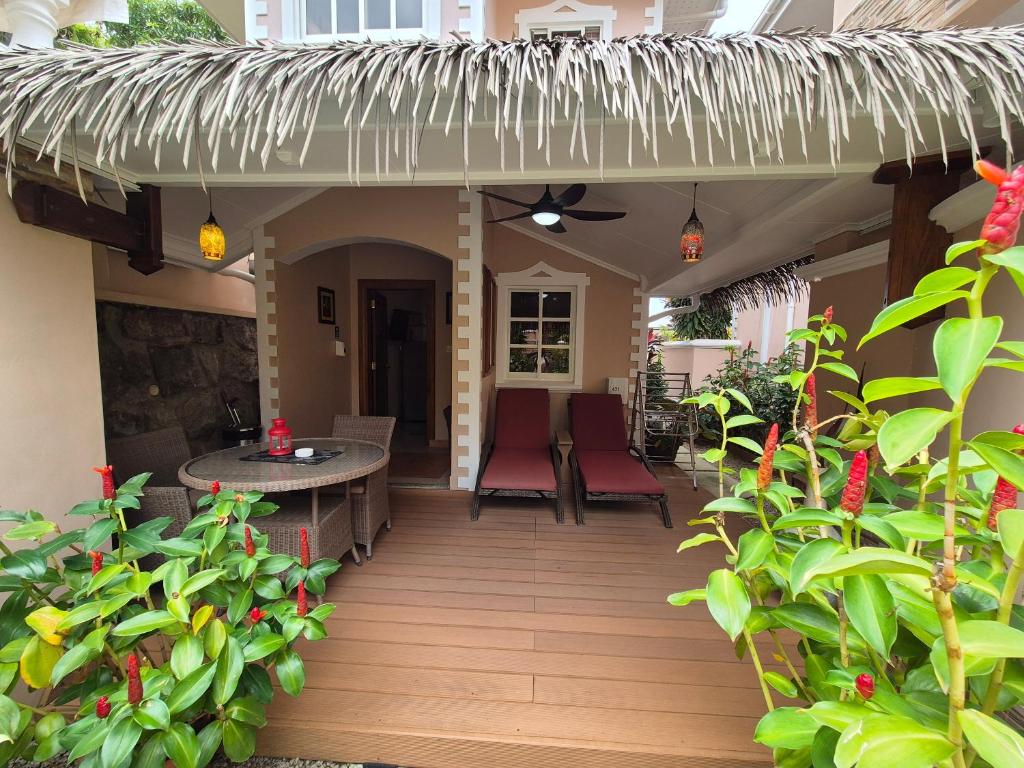 Relaksējoša terase ar divām guļamvietām, galdu un krēsliem, ko ieskauj krāsaini ziedi un zaļa veģetācija. Mājas ieejas apdare ar siltu apgaismojumu un jumtu no salmiem.