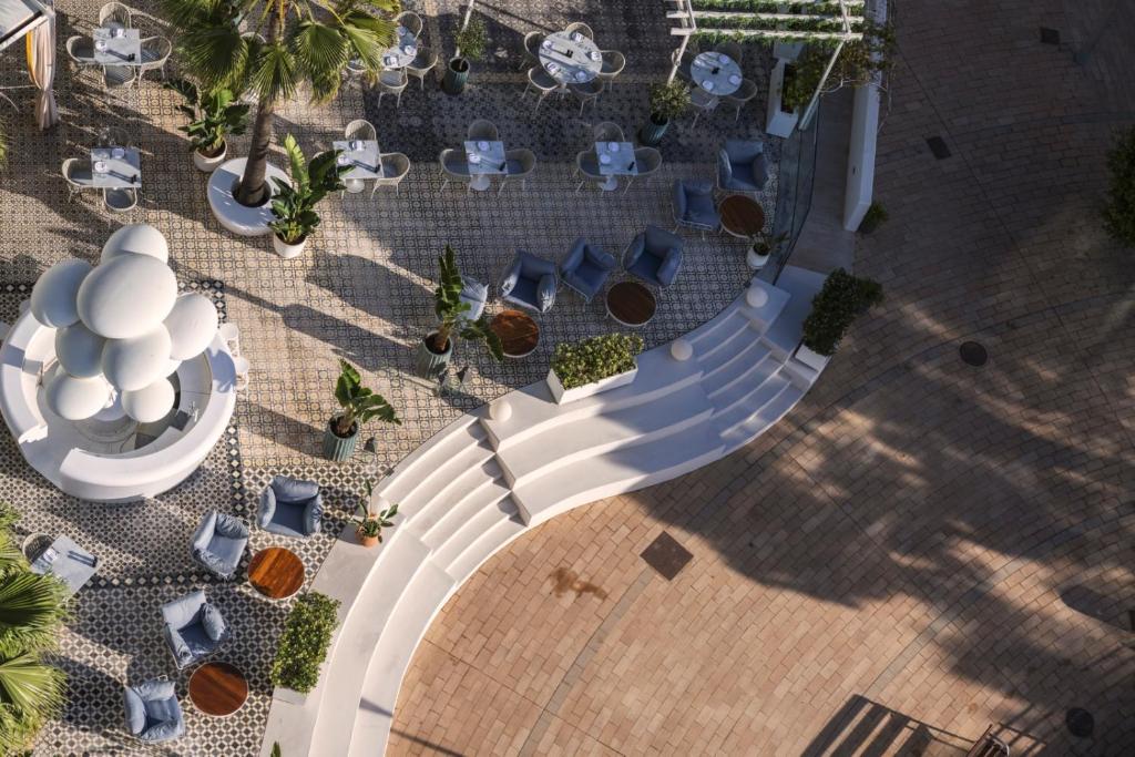 Mūsdienīgs āra kafejnīcas vai restorāna skats, kurā redzami eleganti galdi un krēsli, zaļas palmas un stilizēta strūklaka, kas rada relaksējošu atmosfēru.