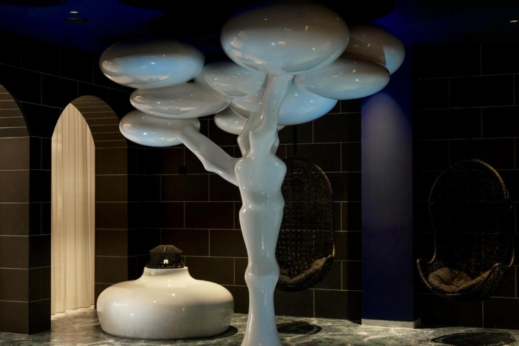 Mūsdienīga mākslas instalācija, kas attēlo stilizētu koku ar gludām, baltām formām un cilvēka figūru, izceļas melnā un tumši zilā interjerā.