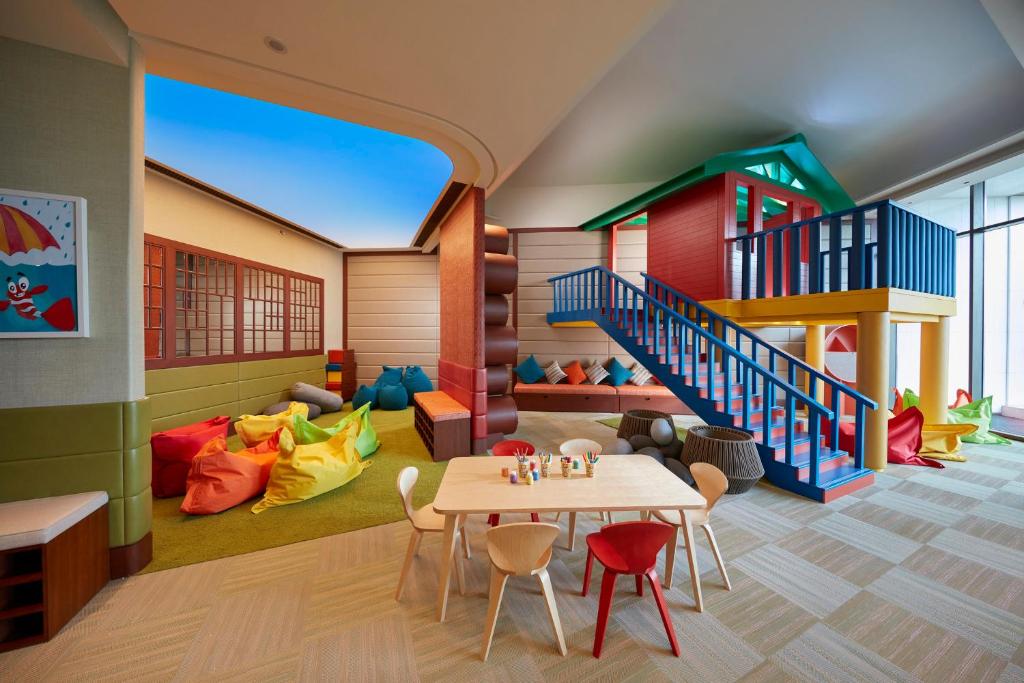 Krāsains bērnu rotaļu istaba ar rotaļu māju, mīkstiem krēsliem un galdu, kas aprīkots ar krāsainām zīmēšanas piederumiem. Istabā ir daudz spilgtu krāsu un komfortablu sēdvietu.