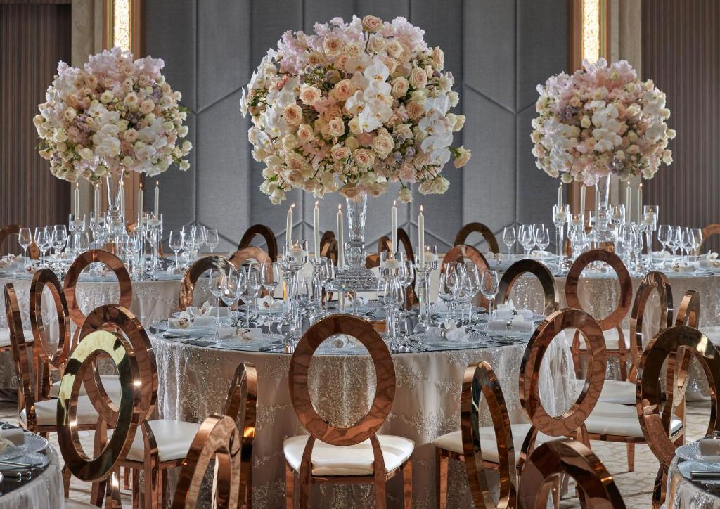 Elegants svinību galds ar greznu ziedu kompozīciju un sudraba traukiem, radot izsmalcinātu atmosfēru. Galds ir rotāts ar stikla svečturiem un modernām krēsliem ar apļveida detaļām.