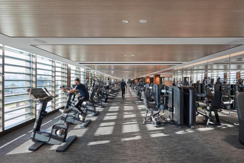 Mūsdienīgs sporta zāles interjers ar daudzām trenažieru iekārtām un lieliem logiem, kas nodrošina dabīgu apgaismojumu. Sportotāji izmanto dažādas ierīces, lai uzlabotu fizisko sagatavotību.