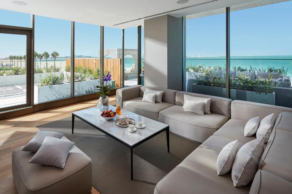 Moderns viesistabas interjers ar plašiem logiem, kas sniedz skatu uz jūru un zaļām terasēm. Uz galda ir augļi un dzērieni, radot relaksējošu atmosfēru.