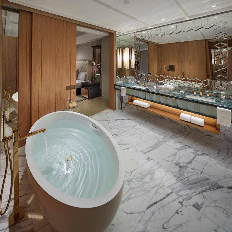 Luksusīga vannas istaba ar ovālu vannu, marmora grīdu un mūsdienīgu dizainu. Interjerā ir koka akcenti un eleganta izlietne ar spoguļiem.