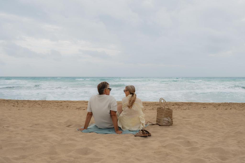 Pāris sēž pludmalē, skatoties uz viļņiem. Apkārt ir smilšaina pludmale un pelēkas debesis.