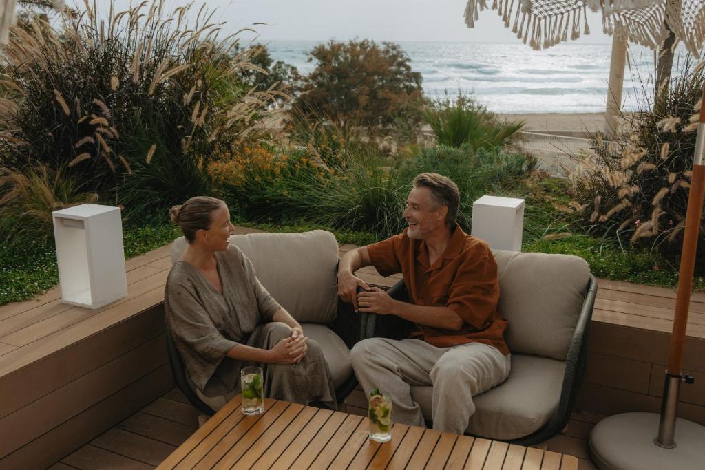 Divi cilvēki relaksējas ārpusē, sēžot dīvānā ar skatu uz jūru, baudot dzērienus un sarunājoties. Apkārt ir zaļumi un moderni apgaismes elementi.