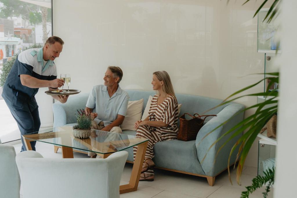 Skaists viesnīcas hols ar viesmīli, kas pasniedz dzērienus diviem viesiem, kas sēž uz dīvāna. Interjers ir moderns un gaišs, radot ērtu atmosfēru.