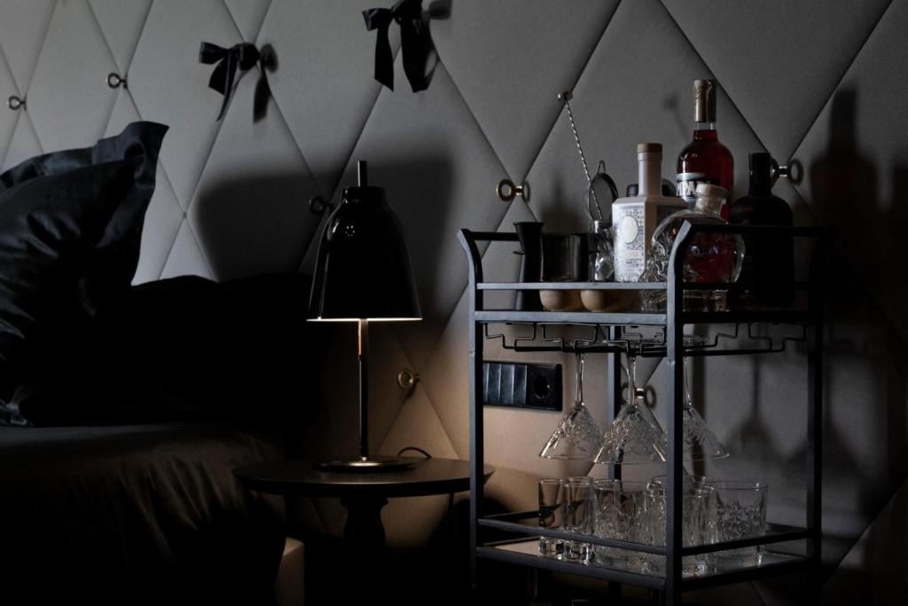 Mūsdienīgs naktsgaldiņš ar melnu lampu un elegantu dzērienu ratiņiem, kas satur dažādas alkohola pudeles un glāzes, pie gultas ar izsmalcinātu sienu apdari.