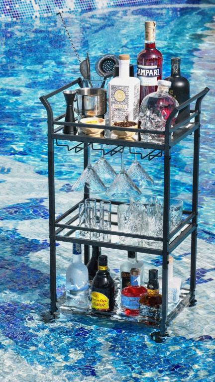 Bāra ratiņi ar dažādiem dzērieniem un glāzēm, novietoti blakus baseinam. Ideāla vide atpūtai un kokteiļu pagatavošanai.