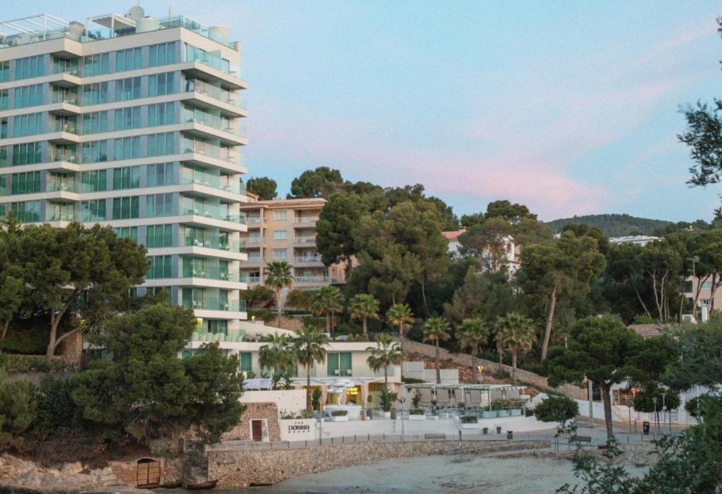 Mūsdienīgs dzīvokļu komplekss ar stikla fasādi, ieskauts zaļām palmām un kokiem, blakus pludmalei. Skats uz apkārtējo ainavu ar kalniem fona.