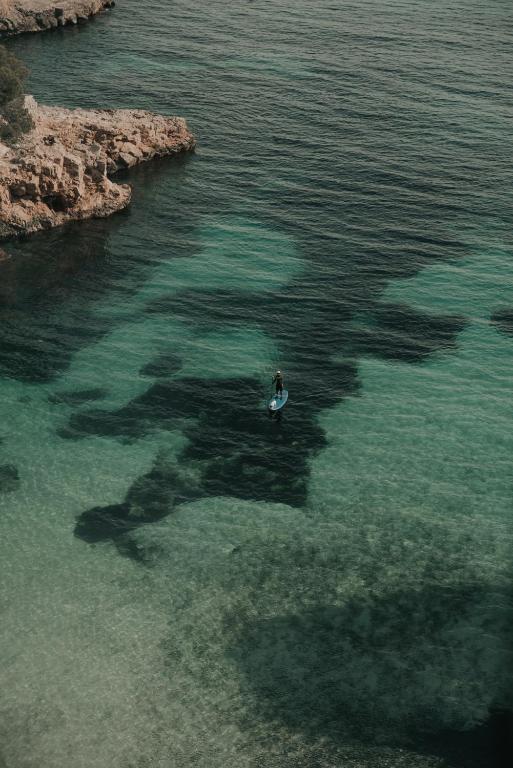 Sērfošana uz stāvā dēļa mierīgā ūdenī, apkārt akmeņains krasts un skaidrs debess.