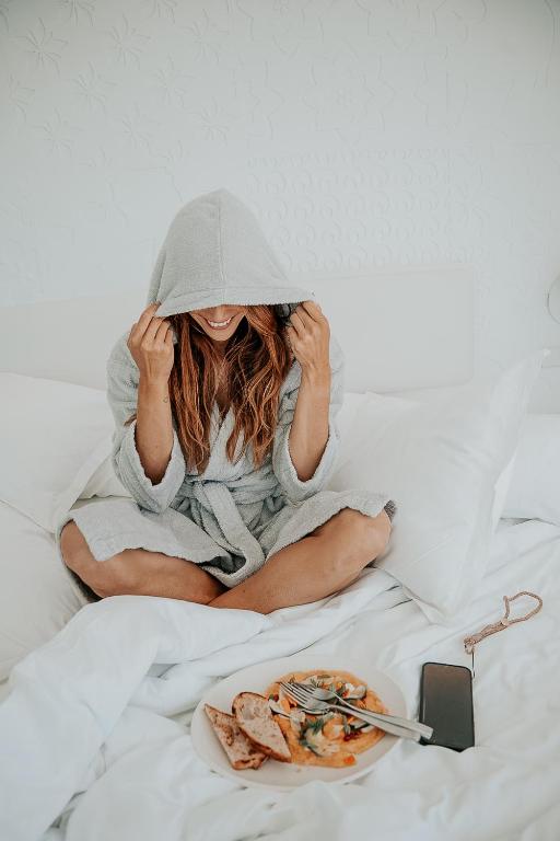 Sieviete ar garām matiem, tērpta pelēkā halātā, sēž gultā, pārklāta ar gultas veļu. Viņa smaida un tur kapuci, blakus ir plates ar ēdienu un telefons.