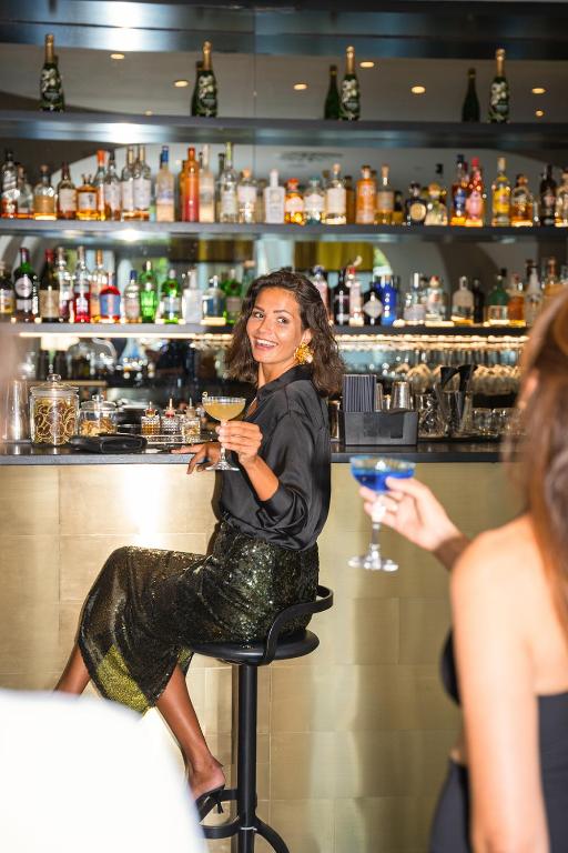 Sieviete sēž pie bāra, turot kokteili, ar smaidu uz sejas un elegantu apģērbu. Fons ir bāra plaukti ar dažādiem dzērieniem.