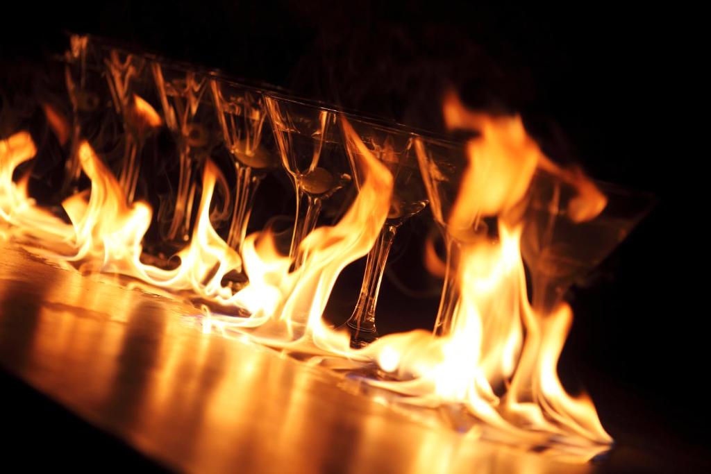 Flambējoši kokteiļu glāzes, kas novietotas rindā, apklātas ar liesmām, radot dramatisku un pievilcīgu skatu.