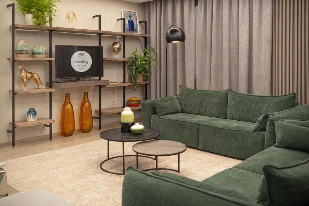 Mūsdienīgs viesnīcas numura interjers ar zaļām dīvānām, stilīgu plauktu un televizoru. Telpā ir pieejami dekoratīvi elementy un augi, radot mājīgu atmosfēru.