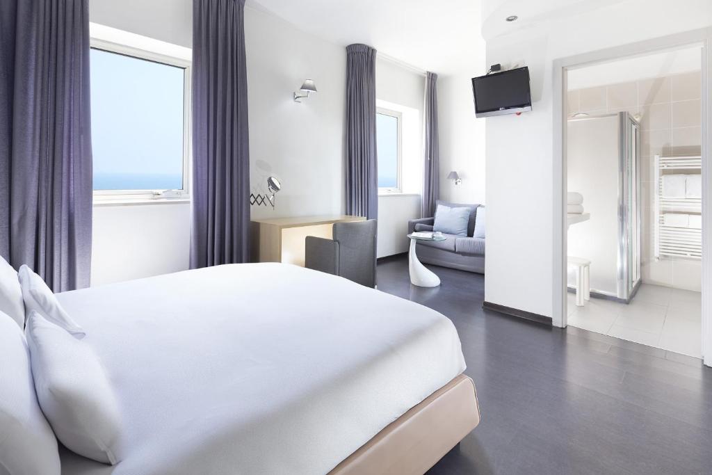 Mūsdienīgs viesnīcas numurs ar gultu, sēžamo stūri un skatu uz jūru. Telpa ir gaiša, ar pelēkām aizkariem un modernām mēbelēm.