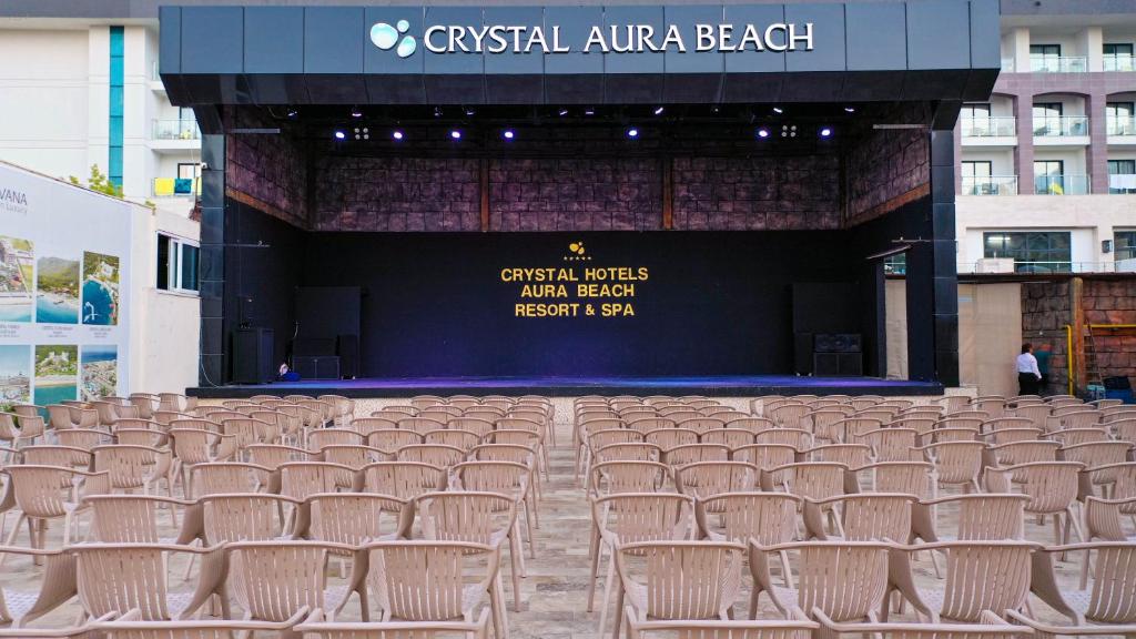 Atklāta skatuve ar krēslu rindām, kas izvietoti priekšā, apgaismota ar prožektoriem. Fons ar nosaukumu "Crystal Aura Beach Resort & Spa".