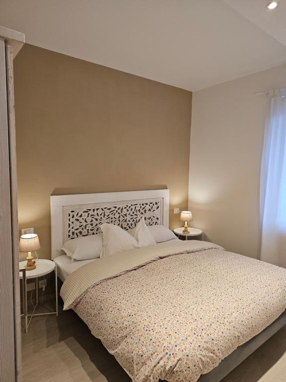 Mūsdienīgs guļamistabas interjers ar baltu gultu un dekoratīvu galvgali, blakus ir divi naktsgaldiņi ar lampām. Siltas krāsas sienas un dabisks apgaismojums rada mājīgu atmosfēru.