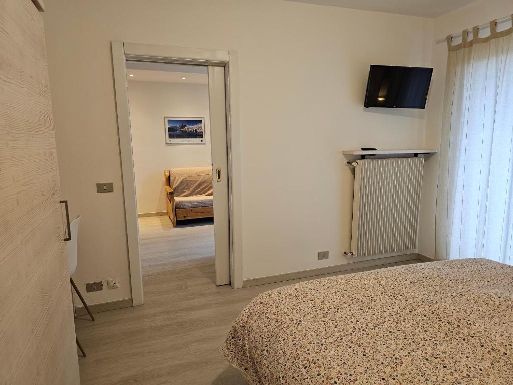 Mūsdienīgs guļamistabas interjers ar gultu, televizoru un lieliem logiem, kas nodrošina dabisko apgaismojumu. Otrā telpā redzama dīvāna zona un ainava uz sienas.