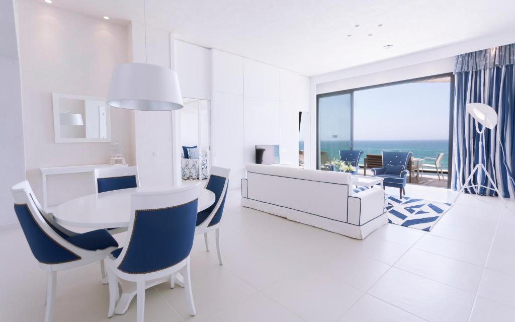 Mūsdienīga viesistaba ar jūras skatu, kurā ir baltas un zilas mēbeles, apļa galdiņš un elegantas krēsli. Gaismas un plašuma sajūta, kas veido relaksējošu atmosfēru.