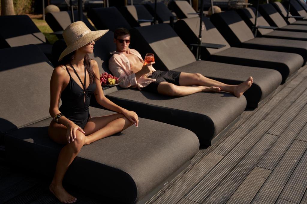 Atpūtas brīdis pie baseina ar diviem jauniešiem, kuri relaksējas uz sauļošanās krēsliem. Viens no viņiem baudīja dzērienu, bet otra persona ir ģērbusies peldkostīmā un uzvelk platmali.
