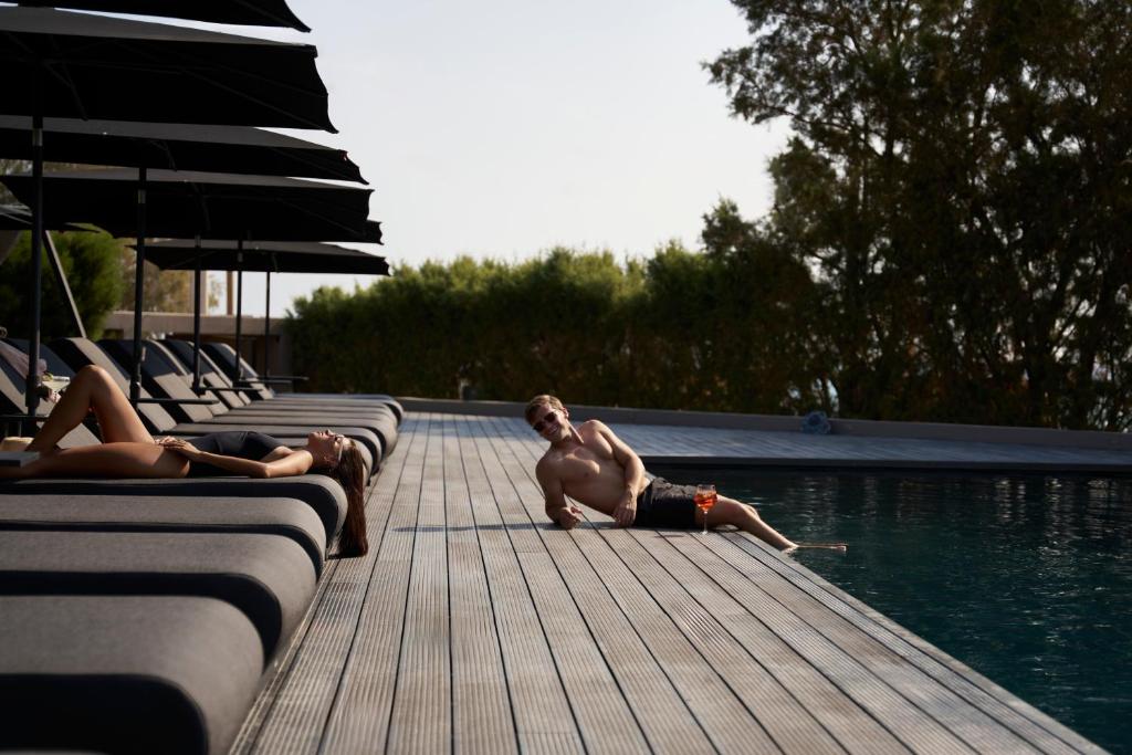 Atpūtas brīvdienas pie baseina ar diviem cilvēkiem, kas relaksējas saulē. Fons ar palmām un saules krēsliem papildina kluso atmosfēru.