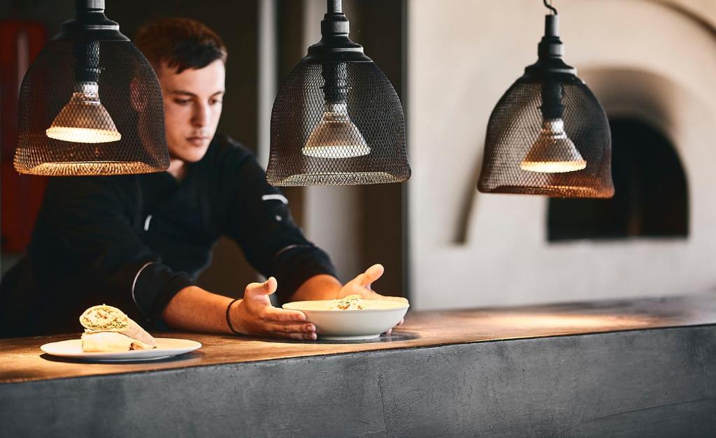 Šefpavārs pasniedz ēdienu elegantā restorānā, izmantojot modernu apgaismojumu un stilīgu interjeru. Uz galda redzami divi šķīvji ar ēdieniem.