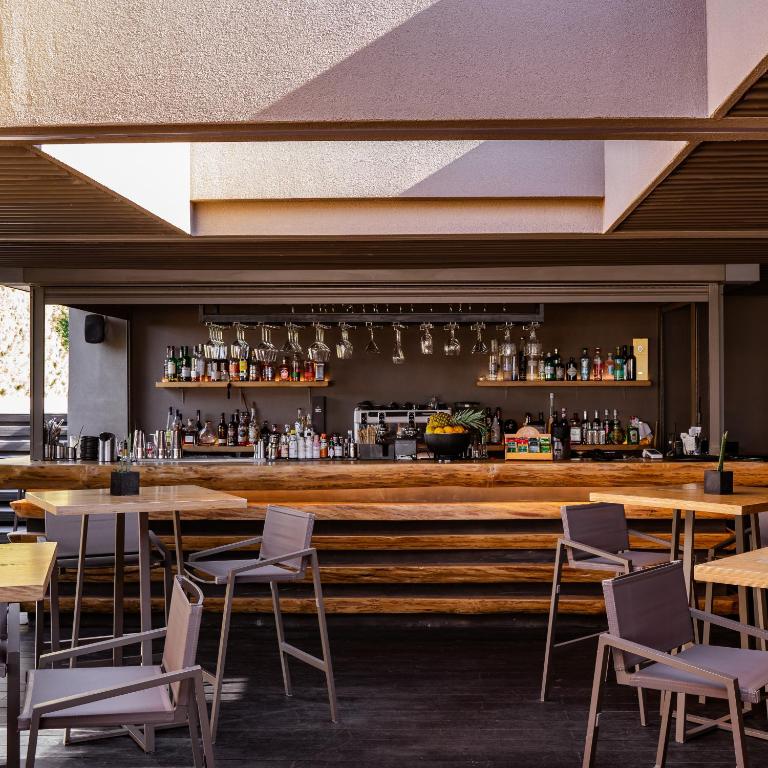 Mūsdienīgs bārs ar koka letes un elegantu interjeru, kurā redzami dažādi alkoholiskie dzērieni un glāzes. Telpa ir gaiša un atvērta, radot relaksējošu atmosfēru.