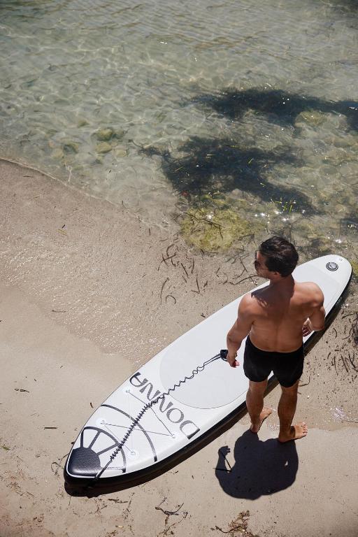 Vīrietis stāv uz pludmales ar sērfošanas dēli, skatoties uz skaidro ūdeni. Apgaismojums ir saulains, radot relaksējošu atmosfēru.