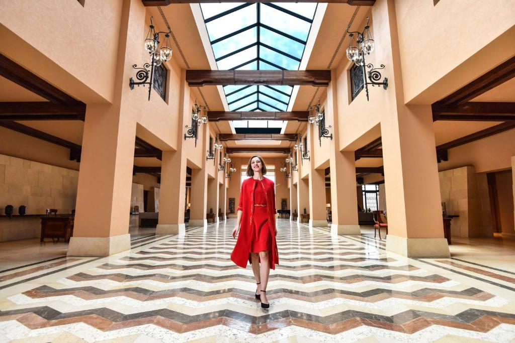 Sieviete sarkana kleitā staigā elegantā viesnīcas foajē ar plašiem logiem un dekoratīvām lampām. Grīda ir veidota ar ģeometriskām rakstām, radot modernu un stilīgu atmosfēru.