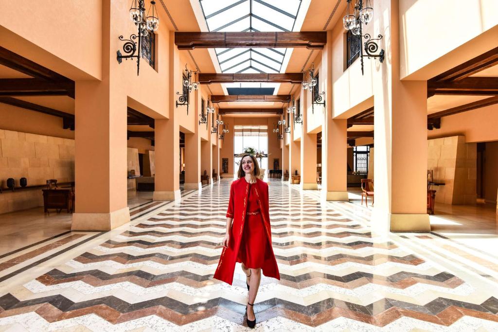 Sieviete sarkanā kleitā stāv modernā un eleganta interjerā ar rakstainu grīdu, kas izceļ plašo telpu. Gaismas caurspīdīgās jumta daļas un stilīgie gaismas ķermeņi piešķir atmosfēru.