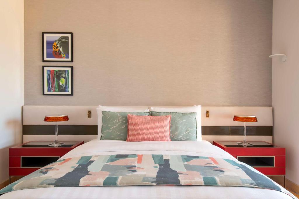 Mūsdienīga guļamistaba ar stilīgu gultu, krāsainām gultas segām un mākslas darbiem uz sienas, radot ērtu un estētisku atmosfēru.