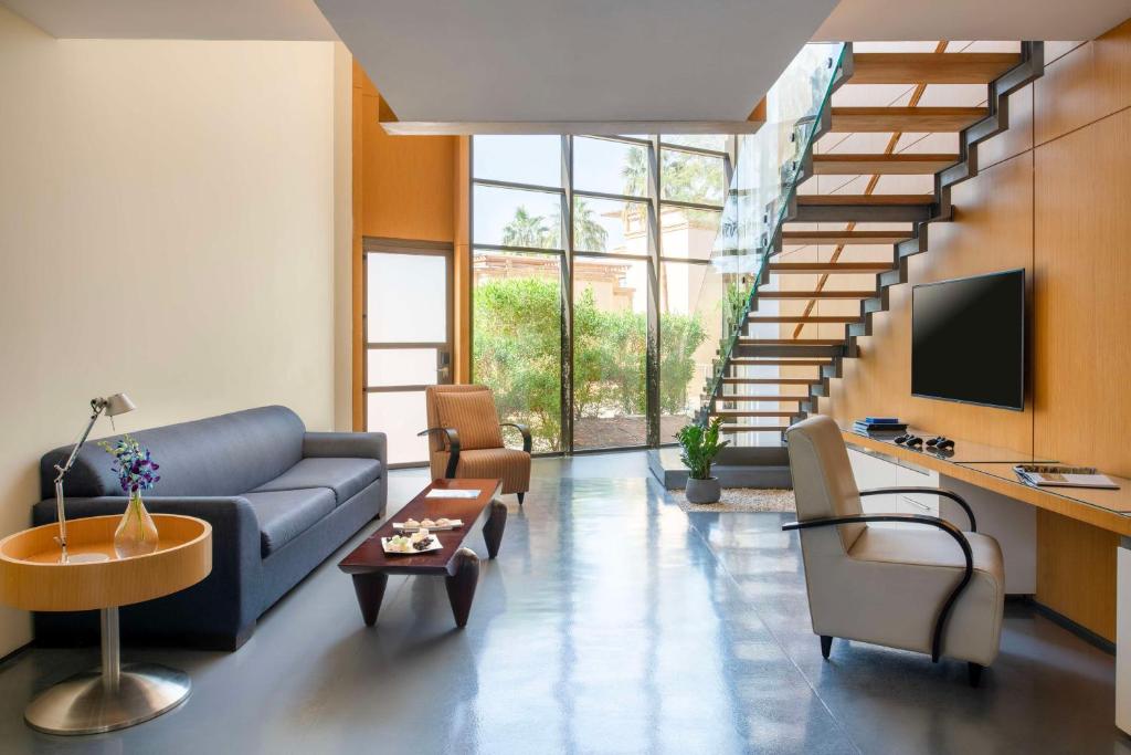 Mūsdienīgs viesistabas interjers ar ērtu dīvānu, koka kāpnēm un lieliem logiem, caur kuriem ieplūst dabiska gaisma. Elegants dizains ar augu elementiem un modernām mēbelēm.