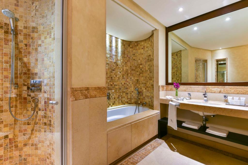 Elegantā vannas istaba ar mozaīkas flīzēm, dušu un vannu, kas nodrošina relaksējošu vidi. Uz galda atrodas higiēnas piederumi un zieds.