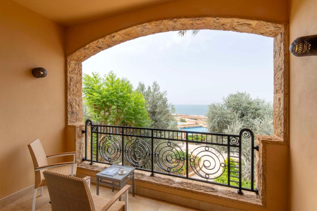 Balkons ar skatu uz jūru un zaļiem kokiem, kur divas krēsli un galds gaida apmeklētājus.