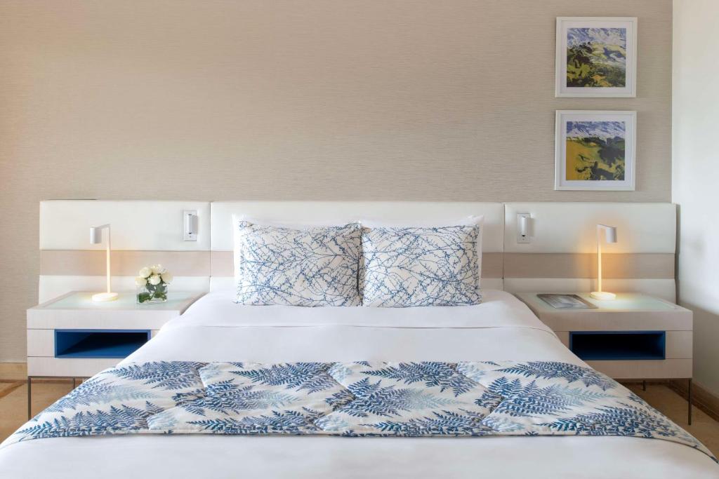 Mūsdienīgs guļamistabas interjers ar gultu, dekorētu ar zilu un balto gultas pārklāju. Blakus gultai atrodas divas naktslampas un neliels ziedu pušķis. Sienās ir divas gleznas ar ainavām.