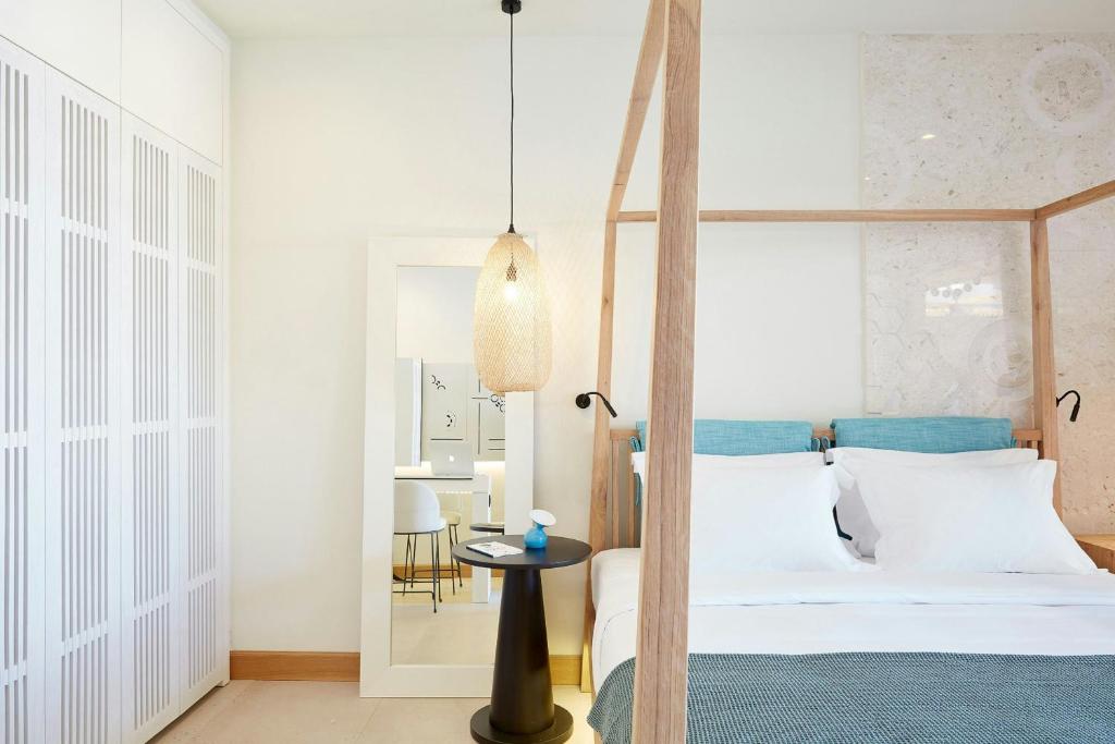 Mūsdienīga guļamistaba ar koka gultu, gaišiem toņiem un elegantu apgaismojumu, kas rada relaksējošu atmosfēru. Redzami arī spogulis un neliela galds ar dekoratīviem elementiem.