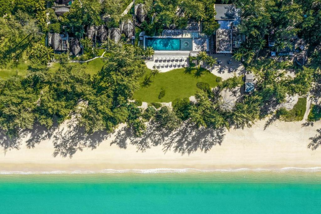 Tuvplāna skats uz pludmali ar smilšainu piekrasti, skaidru zilu ūdeni un viesnīcas kompleksu ar baseinu un zaļām zālēm. Apkārt augoši koki un ēkas rada mierīgu un relaksējošu atmosfēru.