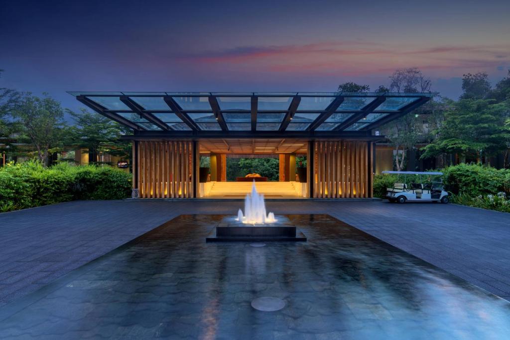 Mūsdienīga viesnīcas ieeja ar stikla jumtu, ūdens strūklaku un apzaļumotu apkārtni, kas rada relaksējošu atmosfēru.